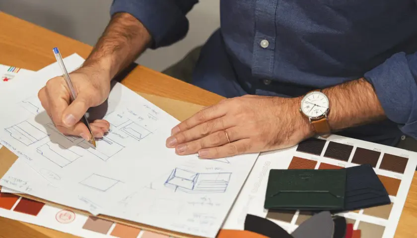 Un designer esquisse des modèles de maroquinerie, entouré d'échantillons de cuir aux teintes variées, évoquant le tannage au chrome.