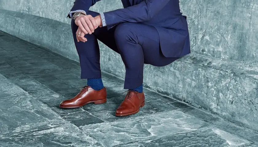 Un homme en costume bleu, avec des chaussures noires et des chaussettes bleues à chevrons, est assis sur un banc en pierre.