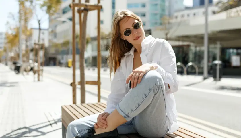Une femme assise sur un banc, portant un chemisier blanc, un jean décontracté et des mocassins noirs. Son outfit simple et chic est parfait pour une journée en ville.