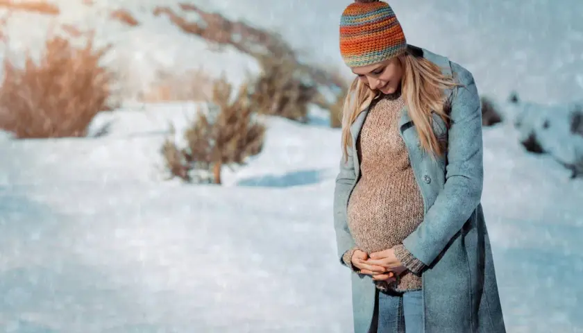 Une femme enceinte, bien emmitouflée dans un manteau et un bonnet coloré, pose dans un paysage enneigé. Elle montre comment s'habiller enceinte en hiver avec style et confort.