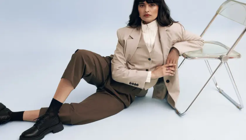 Femme en tenue professionnelle, blazer beige, pantalon marron, et chaussure entretien d'embauche femme noire. Assise près d'une chaise transparente.
