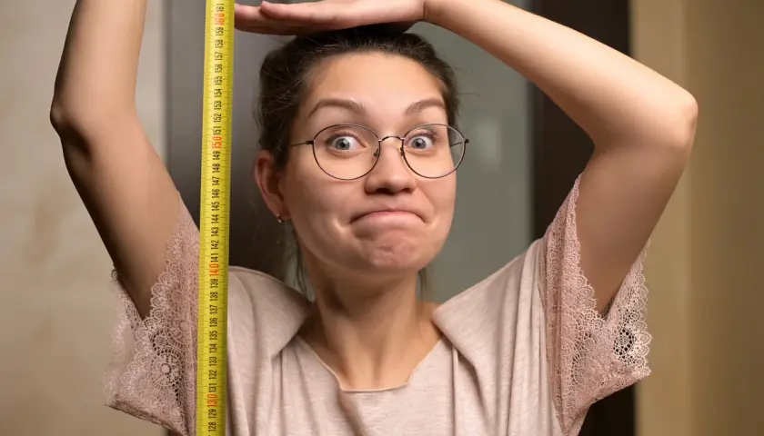Une femme trapue, lunettes rondes, mesure sa taille avec un mètre ruban jaune. Elle a une expression amusée.