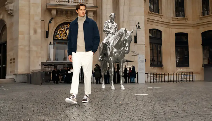 Un homme porte une chaussure avec un chino blanc, un pull à col roulé beige et une veste bleu marine devant une sculpture de cheval argenté.