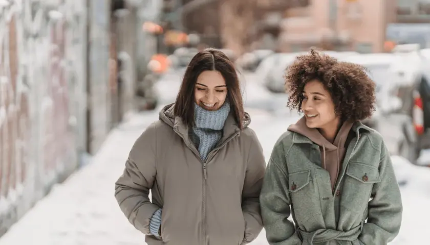 Deux amies discutent en marchant dans la neige. Une idée outfit hiver parfaite : doudoune beige et manteau vert ceinturé.
