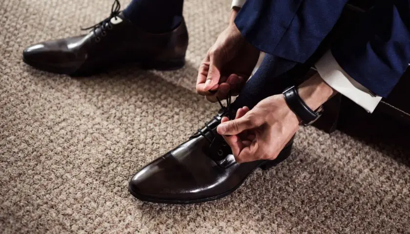 Un homme en costume bleu marine attache ses chaussures vernies marron. Quel chaussure avec un costume pour un look impeccable.