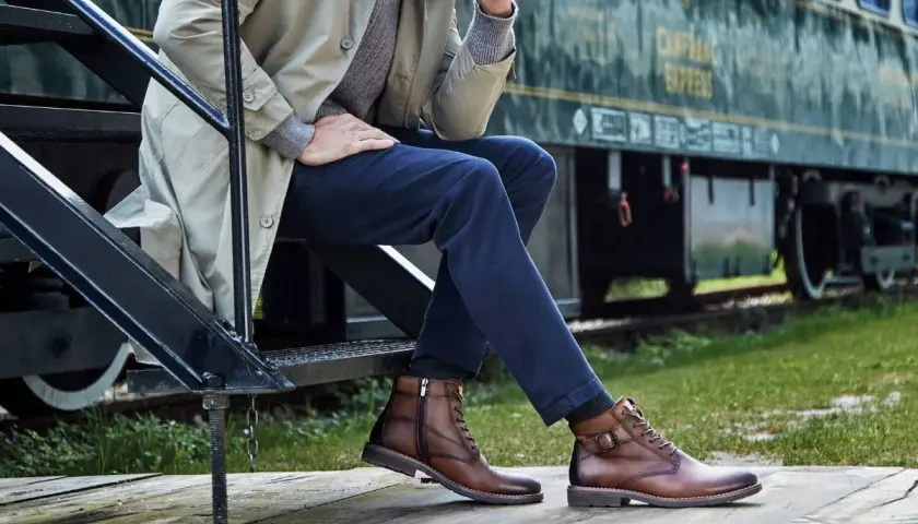 Un homme élégant, assis près d'un train, porte des bottines marron. Une inspiration pour savoir quelles chaussures porter avec un jean homme.