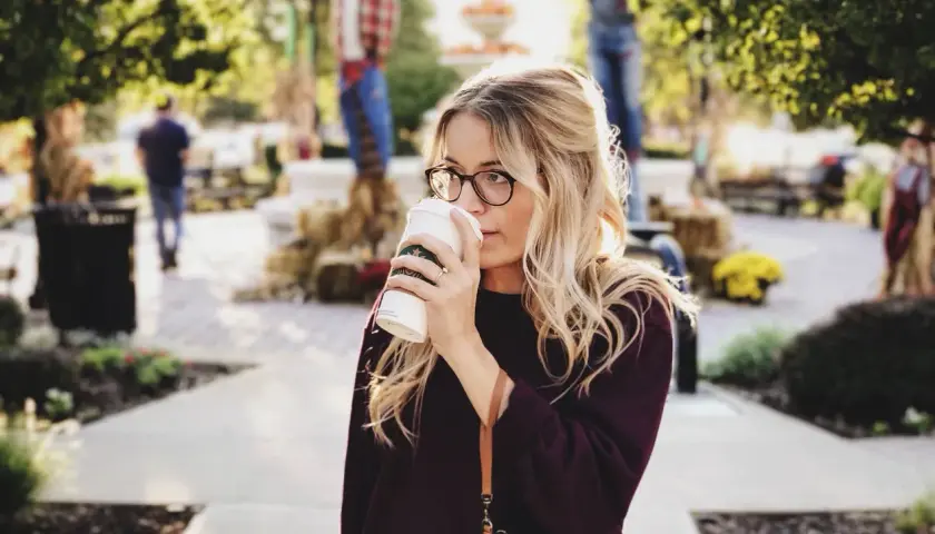 Une femme blonde avec des lunettes boit un café. Elle porte un pull bordeaux. Elle se demande comment dois je m habiller demain.