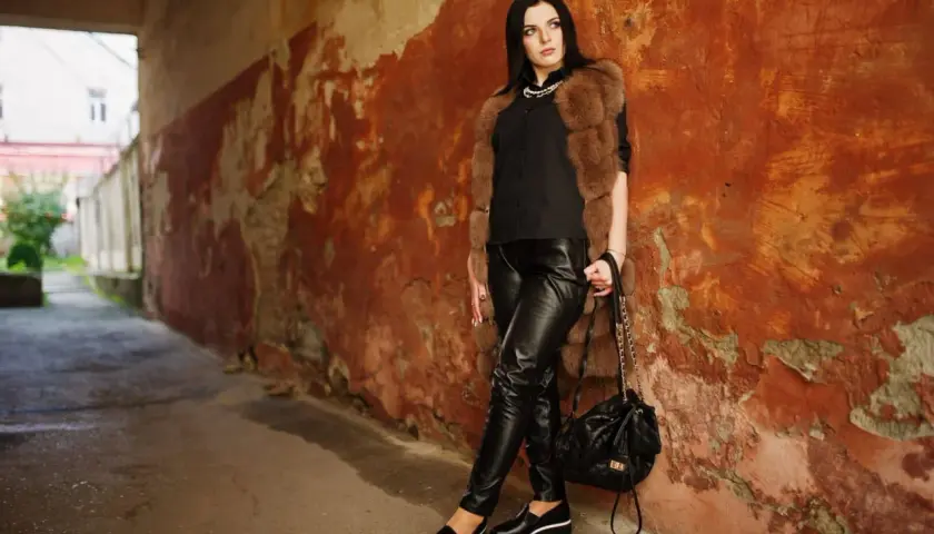 Une femme pose contre un mur décrépit, vêtue d'un pantalon en cuir noir. Apprenez comment nettoyer un pantalon en cuir pour qu'il reste aussi beau.