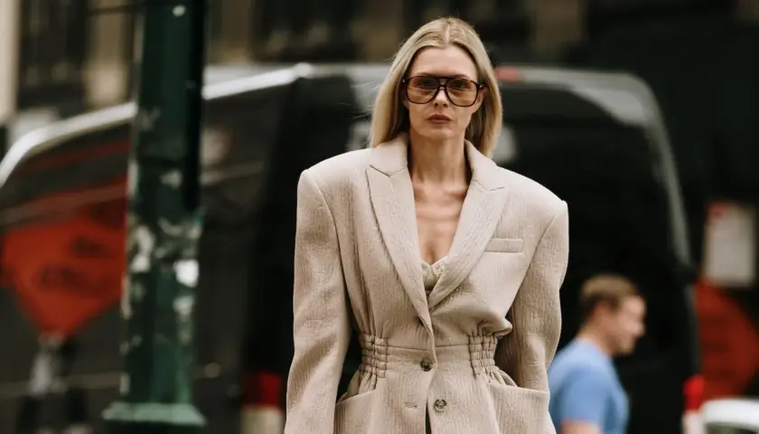 Une femme porte une tenue avec un blazer blanc chic, des lunettes de soleil oversize et un top en dentelle. Elle déambule dans la rue.