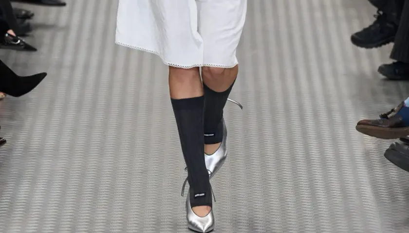 Une mannequin défile avec une tenue blanche et des chaussettes hautes grises, rehaussée par des chaussures argentées pointues.