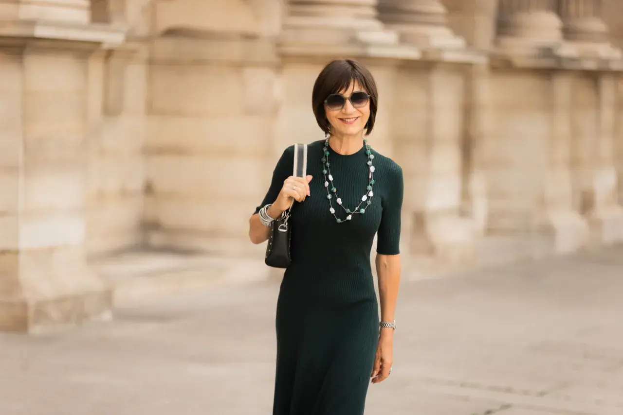 Une femme élégante porte une robe midi vert foncé, un collier de perles et des lunettes de soleil. C'est quoi une robe midi ? C'est une robe qui arrive à mi-mollet.