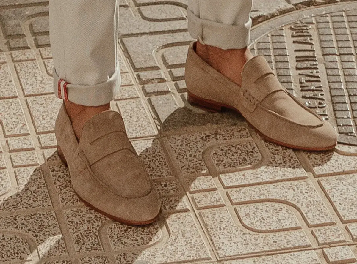 Un homme porte un mocassin à la mode en daim beige, avec un pantalon retroussé, sur un trottoir en dalles.