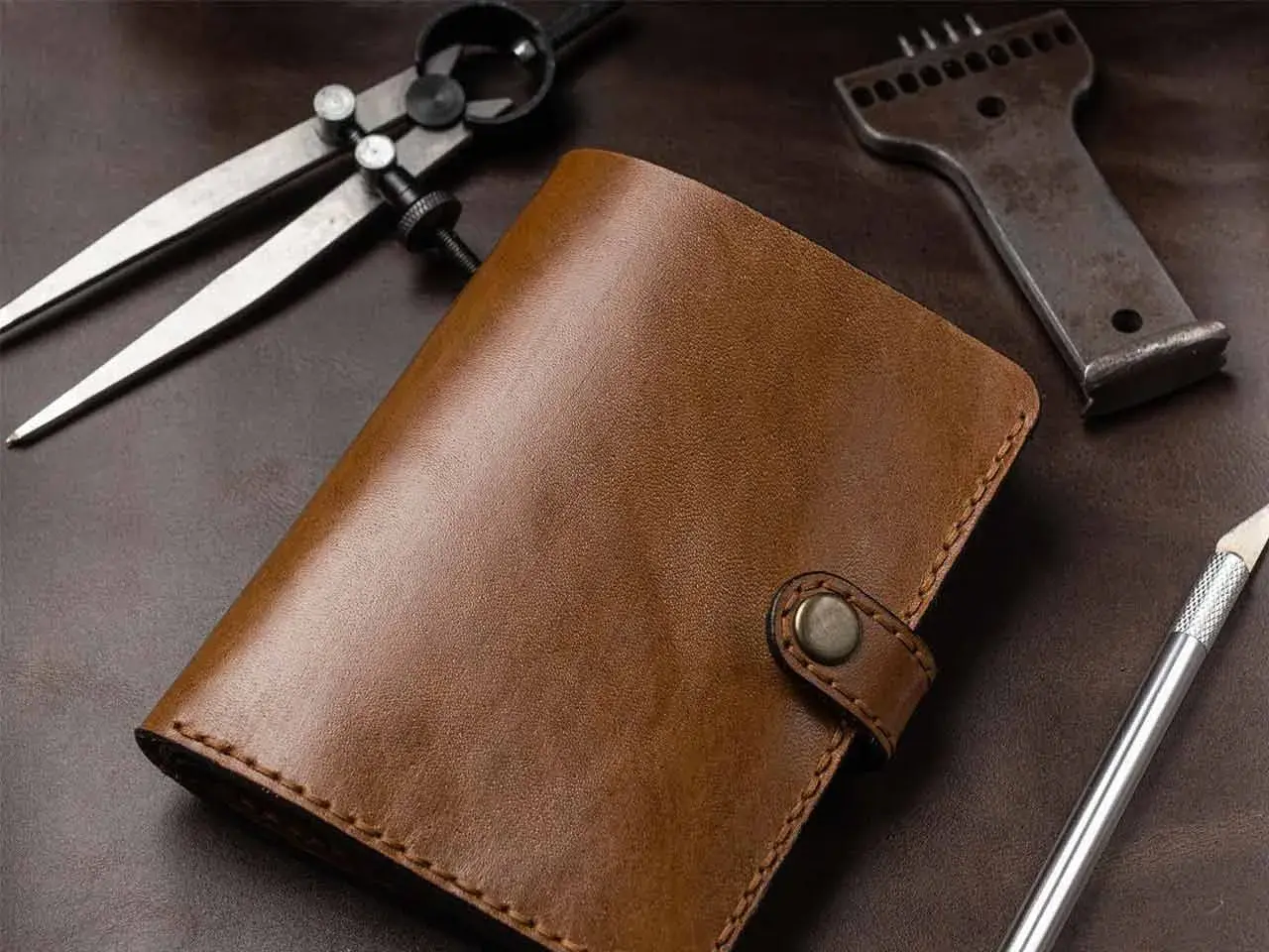 Portefeuille en cuir brun, entouré d'outils de **tannage du cuir** : compas, poinçon et scalpel.
