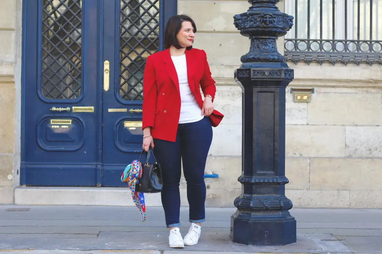Une femme élégante montre comment porter un blazer rouge femme avec un jean et des baskets. Elle tient un sac noir orné d'un foulard coloré.