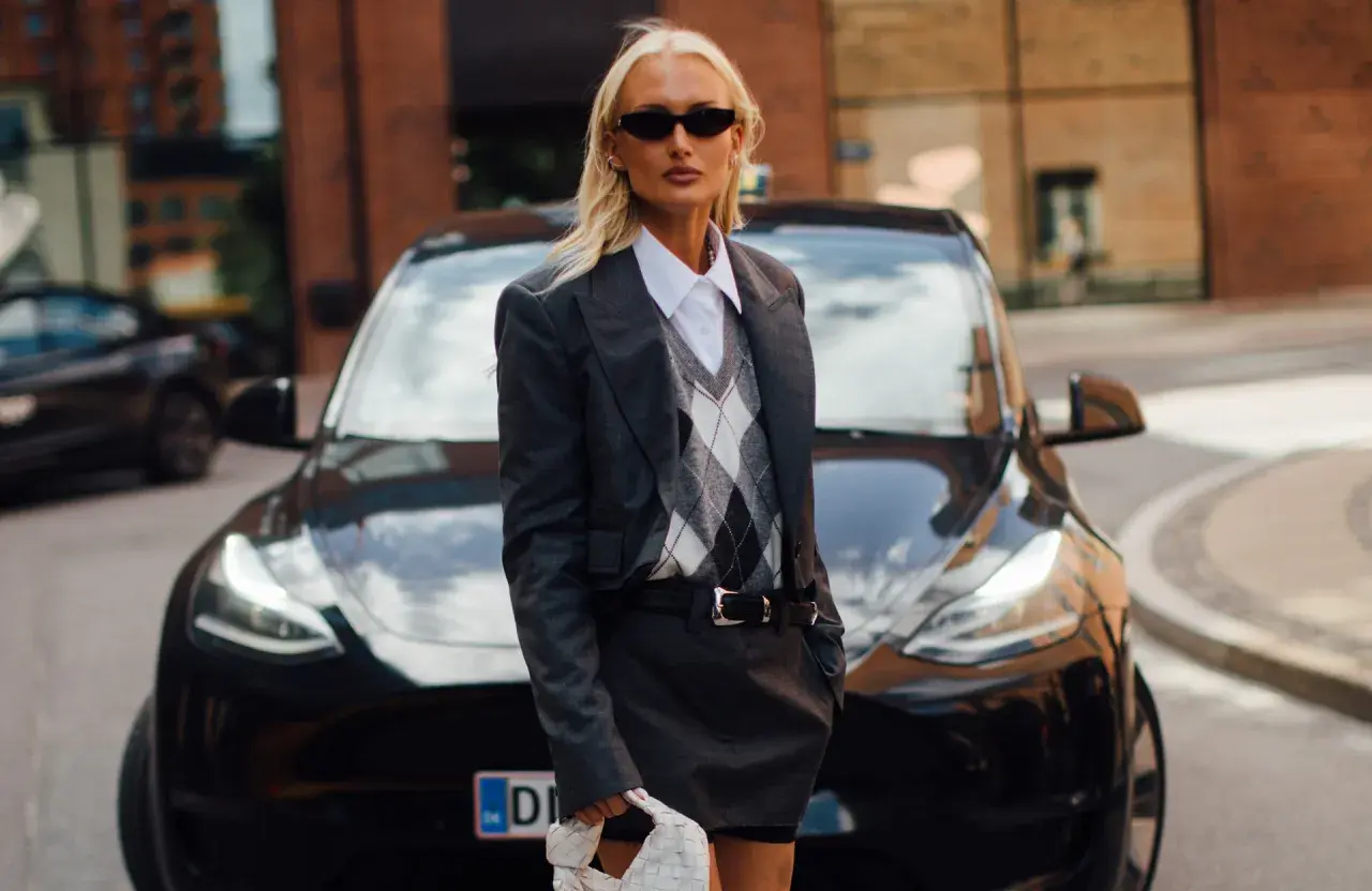 Une femme stylée devant une Tesla noire, portant un ensemble blazer et jupe gris. Un look parfait pour savoir quel blazer pour quelle morphologie.