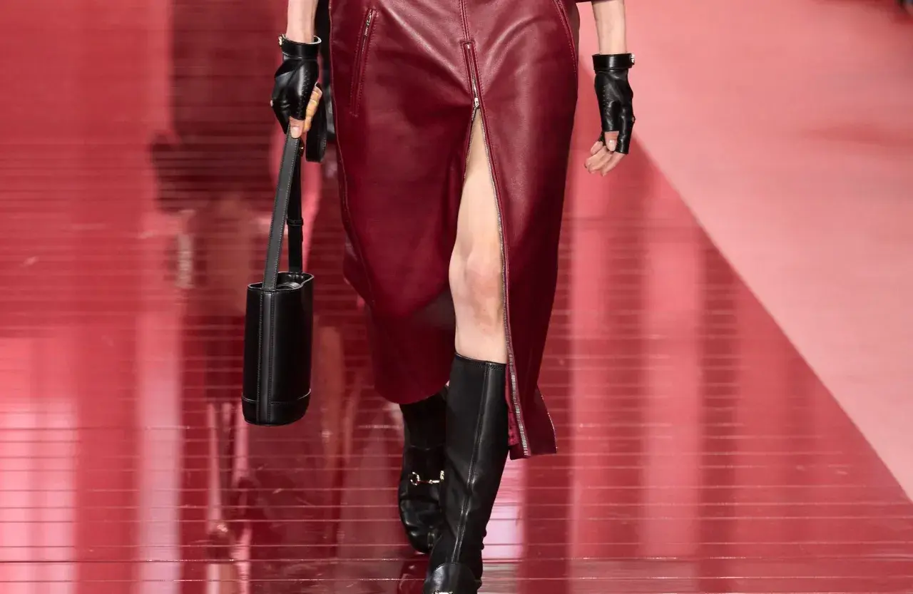 Mannequin portant une jupe courte en cuir bordeaux fendue et des bottines noires. Sac cylindrique noir et gants en cuir.