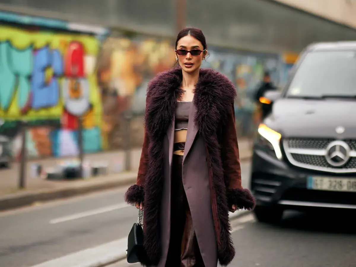 Une femme porte un manteau long en fausse fourrure prune, un exemple de types de manteaux femme tendance. Elle tient un sac noir.