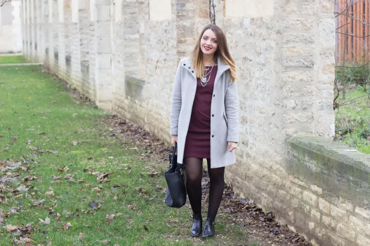 Une femme porte une robe bordeaux et un manteau gris clair. L'association couleur bordeaux vêtements est élégante.