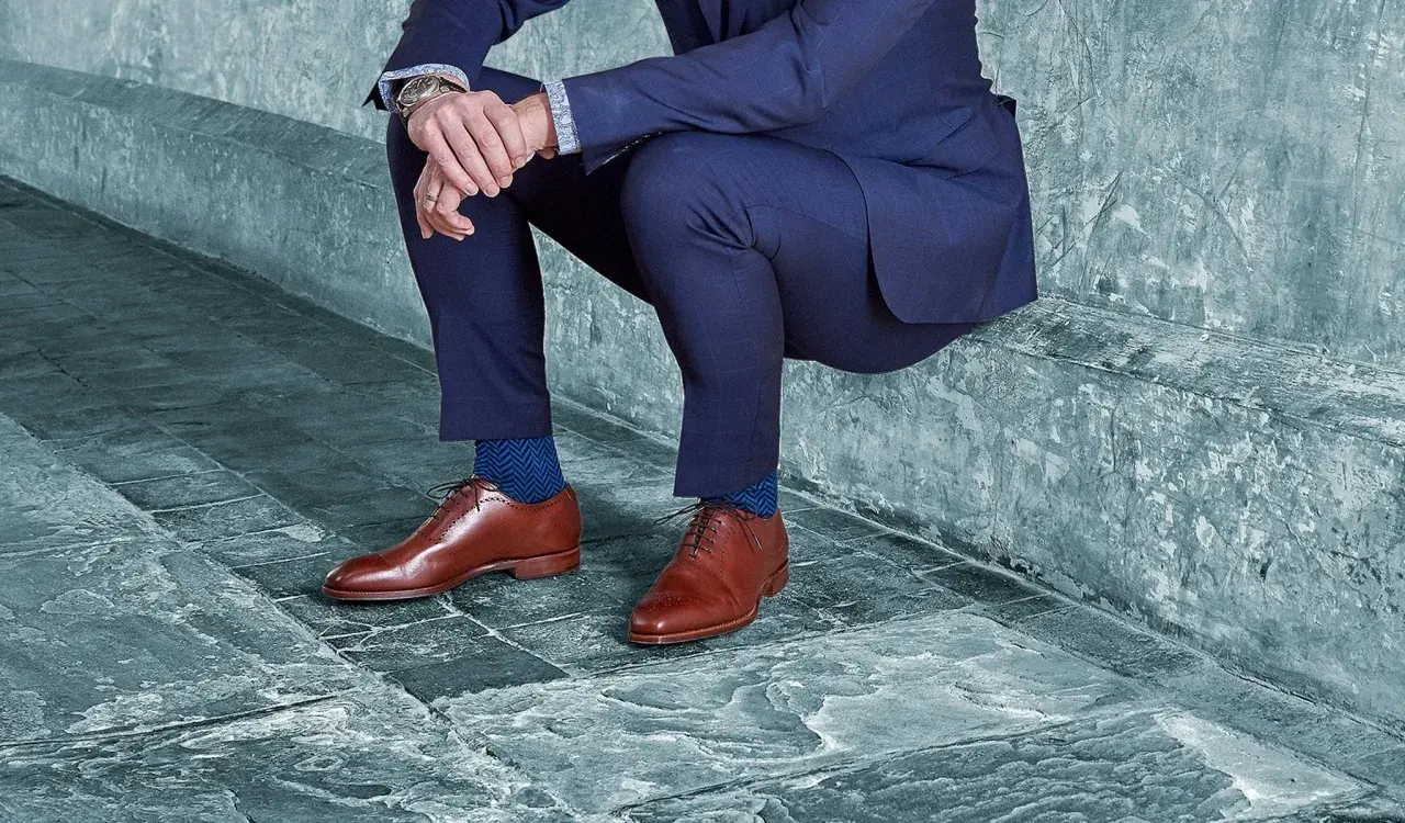 Un homme en costume bleu, avec des chaussures noires et des chaussettes bleues à chevrons, est assis sur un banc en pierre.