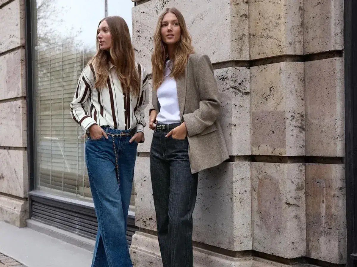 Deux femmes posent devant un mur en pierre. L'une porte un jean tapered femme, une chemise rayée et une ceinture chaîne. L'autre, un blazer et un jean.