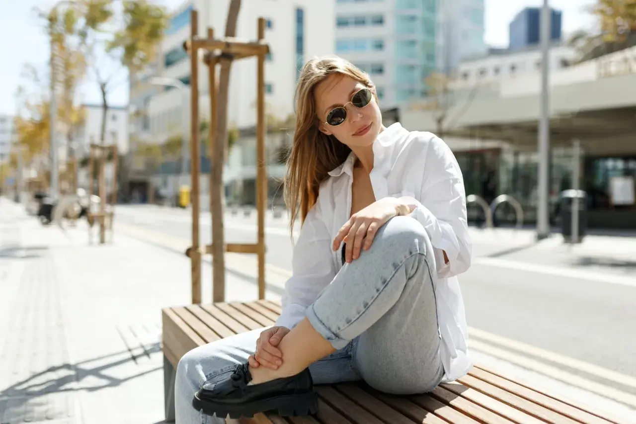 Une femme assise sur un banc, portant un chemisier blanc, un jean décontracté et des mocassins noirs. Son outfit simple et chic est parfait pour une journée en ville.