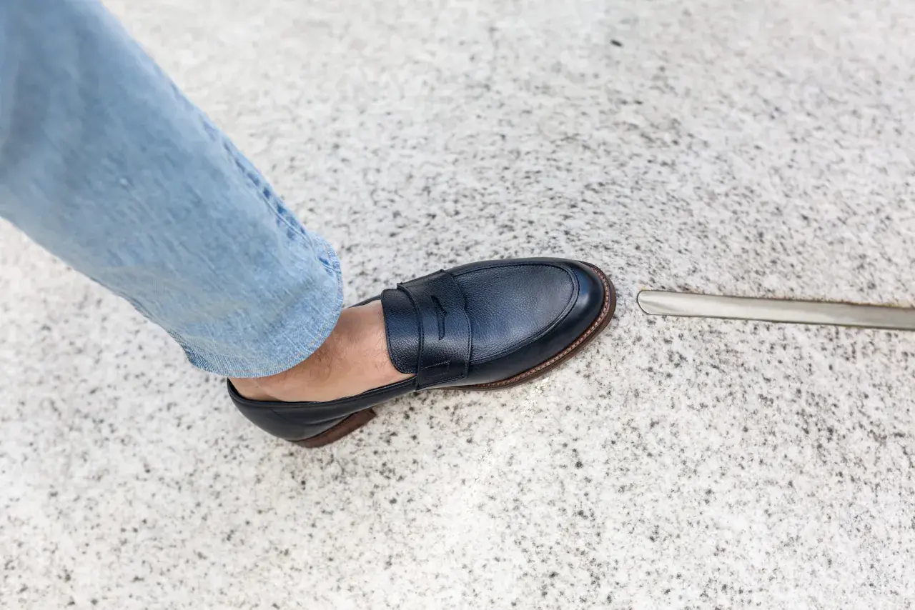 Un pied en jean bleu porte un mocassin bleu marine. La semelle extérieure de la chaussure, signée par un cordonnier, est visible.