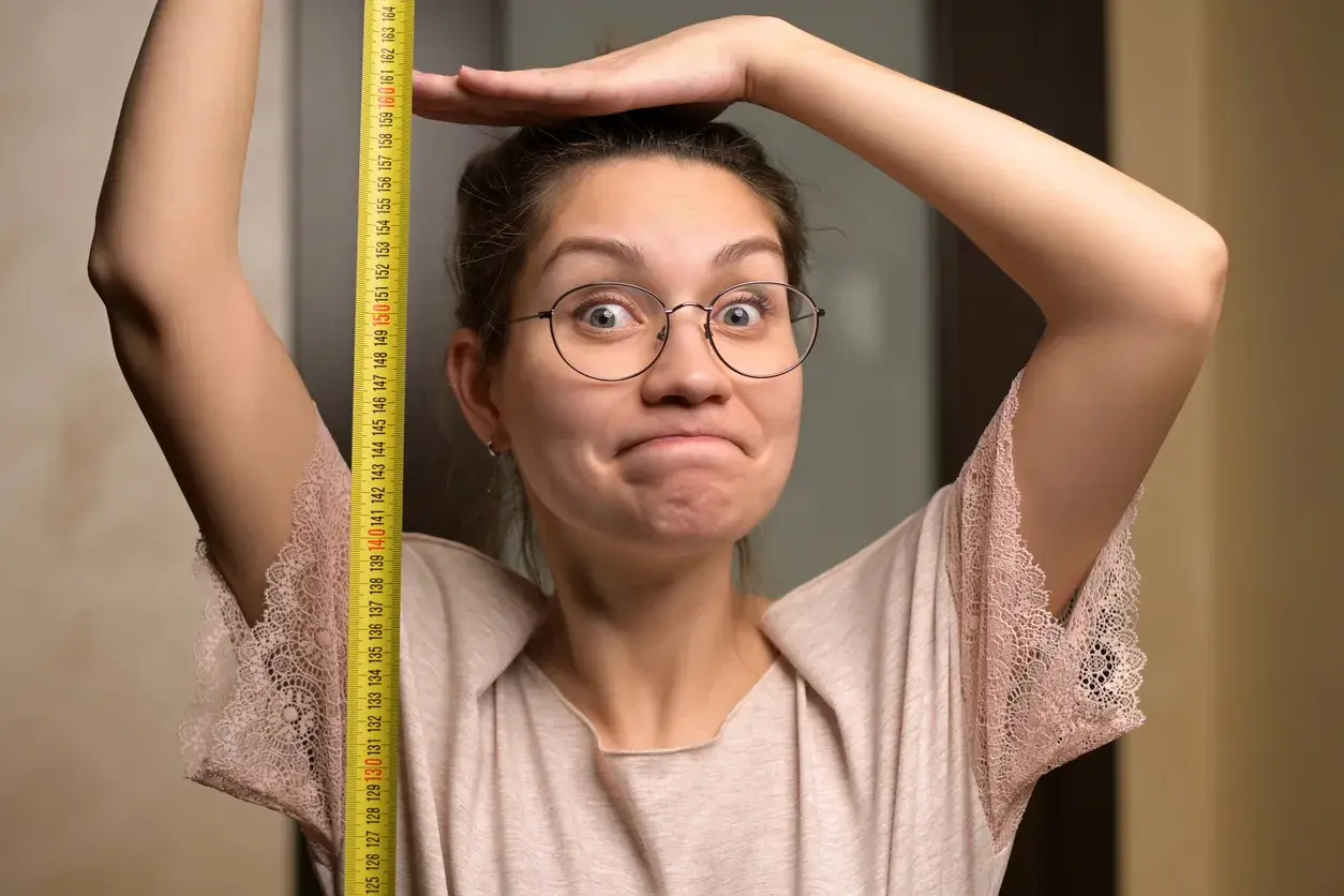 Une femme trapue, lunettes rondes, mesure sa taille avec un mètre ruban jaune. Elle a une expression amusée.