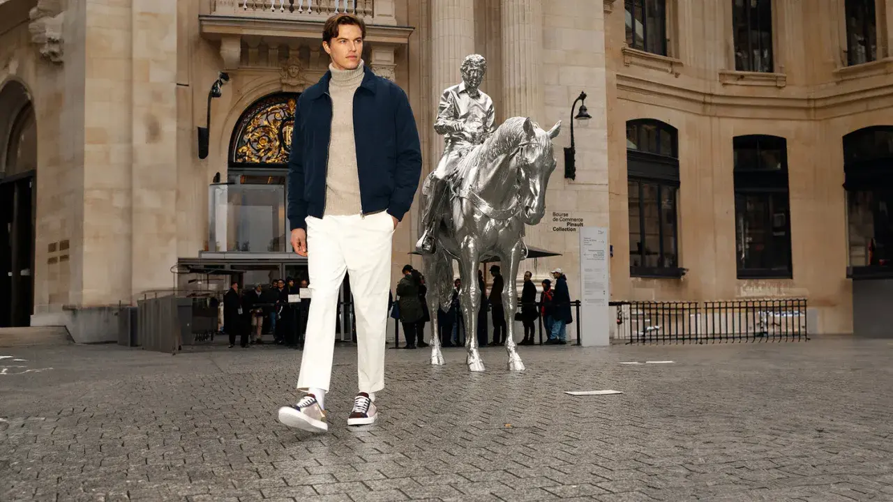 Un homme porte une chaussure avec un chino blanc, un pull à col roulé beige et une veste bleu marine devant une sculpture de cheval argenté.