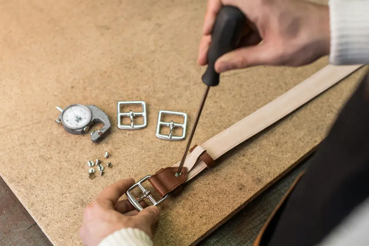 Artisan travaillant sur une ceinture en cuir, utilisant un tournevis pour fixer une boucle. Outils de mesure et boucles supplémentaires à proximité.
