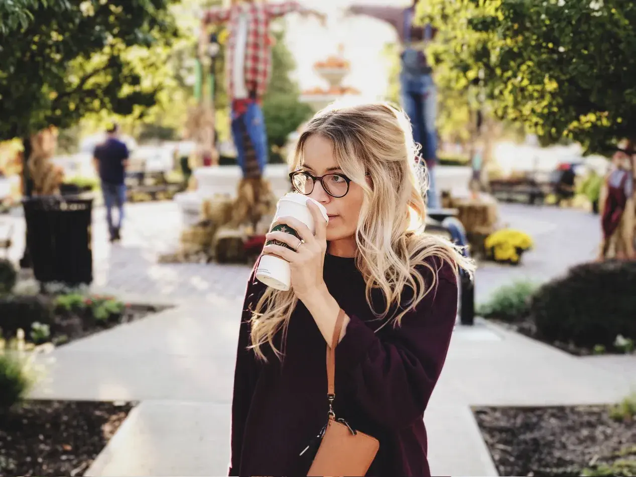 Une femme blonde avec des lunettes boit un café. Elle porte un pull bordeaux. Elle se demande comment dois je m habiller demain.