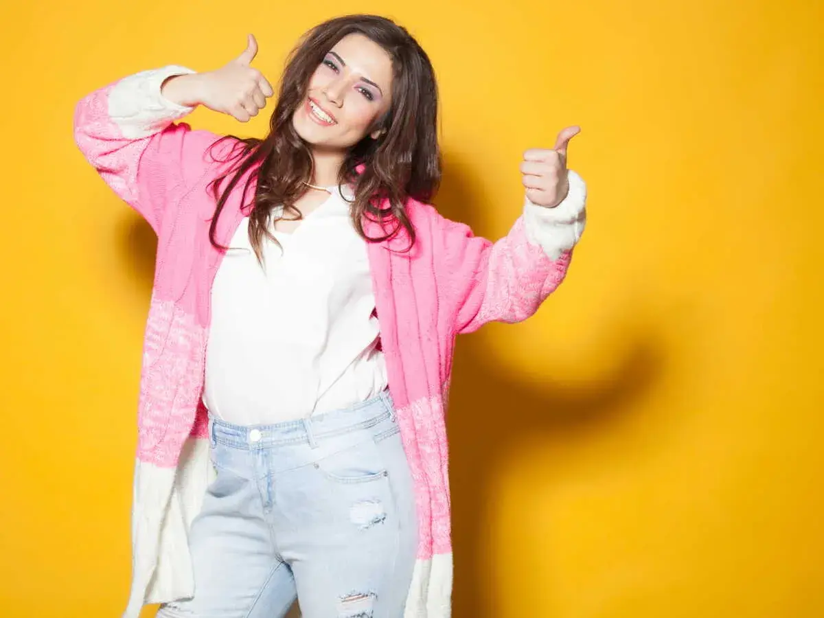 Une femme souriante, petite et ronde, montre comment s'habiller avec un gilet rose et un jean. Elle fait un signe de pouce levé.