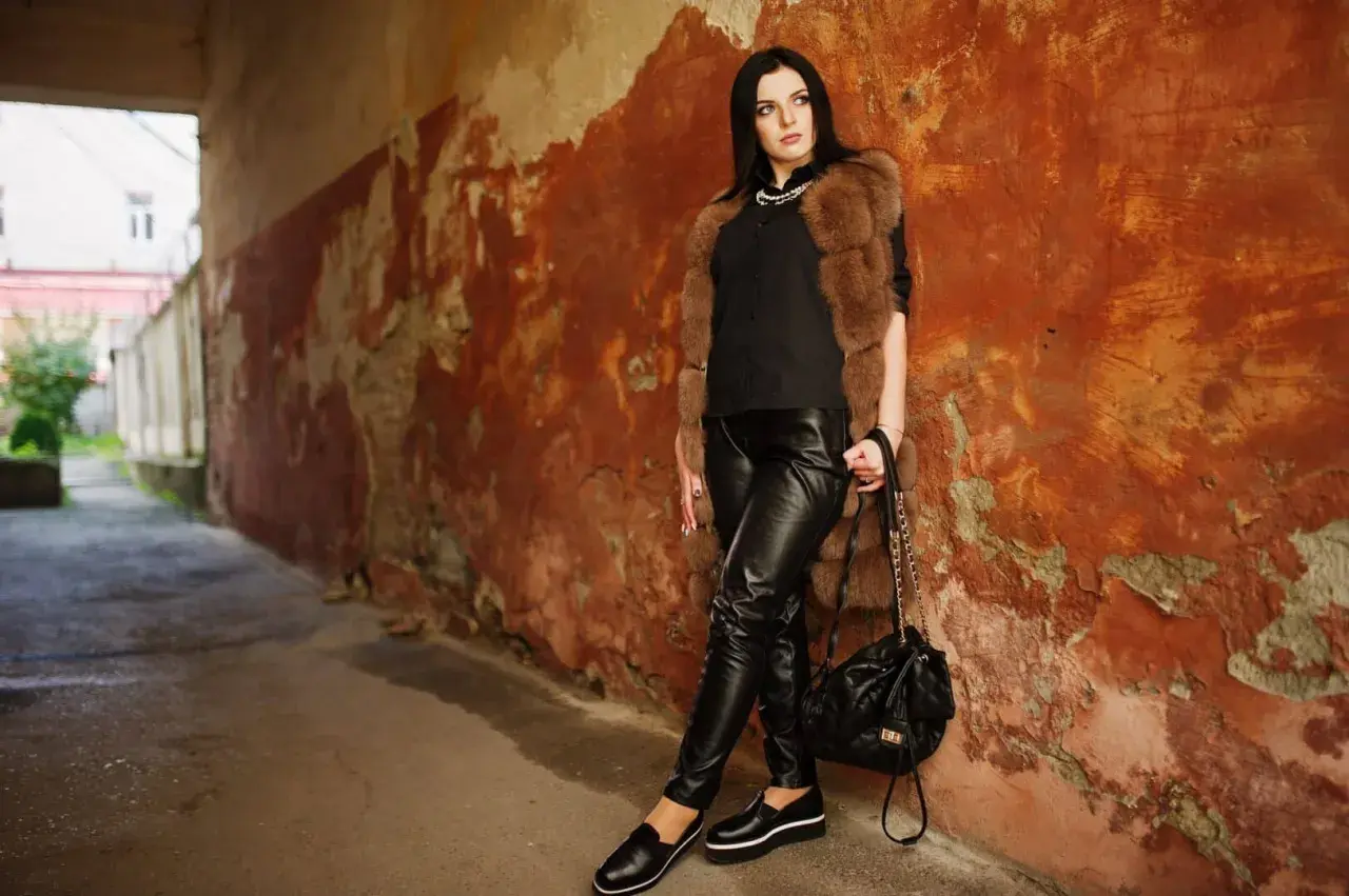 Une femme pose contre un mur décrépit, vêtue d'un pantalon en cuir noir. Apprenez comment nettoyer un pantalon en cuir pour qu'il reste aussi beau.