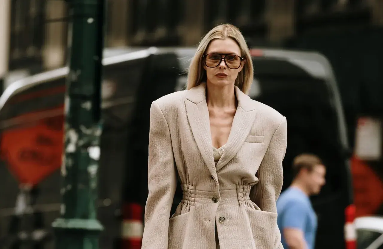 Une femme porte une tenue avec un blazer blanc chic, des lunettes de soleil oversize et un top en dentelle. Elle déambule dans la rue.