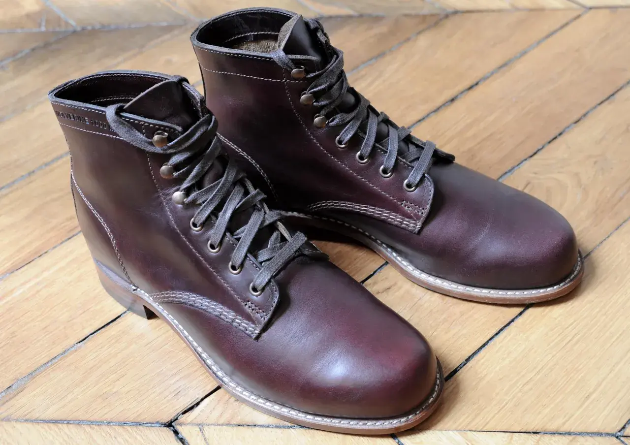 Bottes marron en cuir gras, lacets gris, sur un parquet en bois.