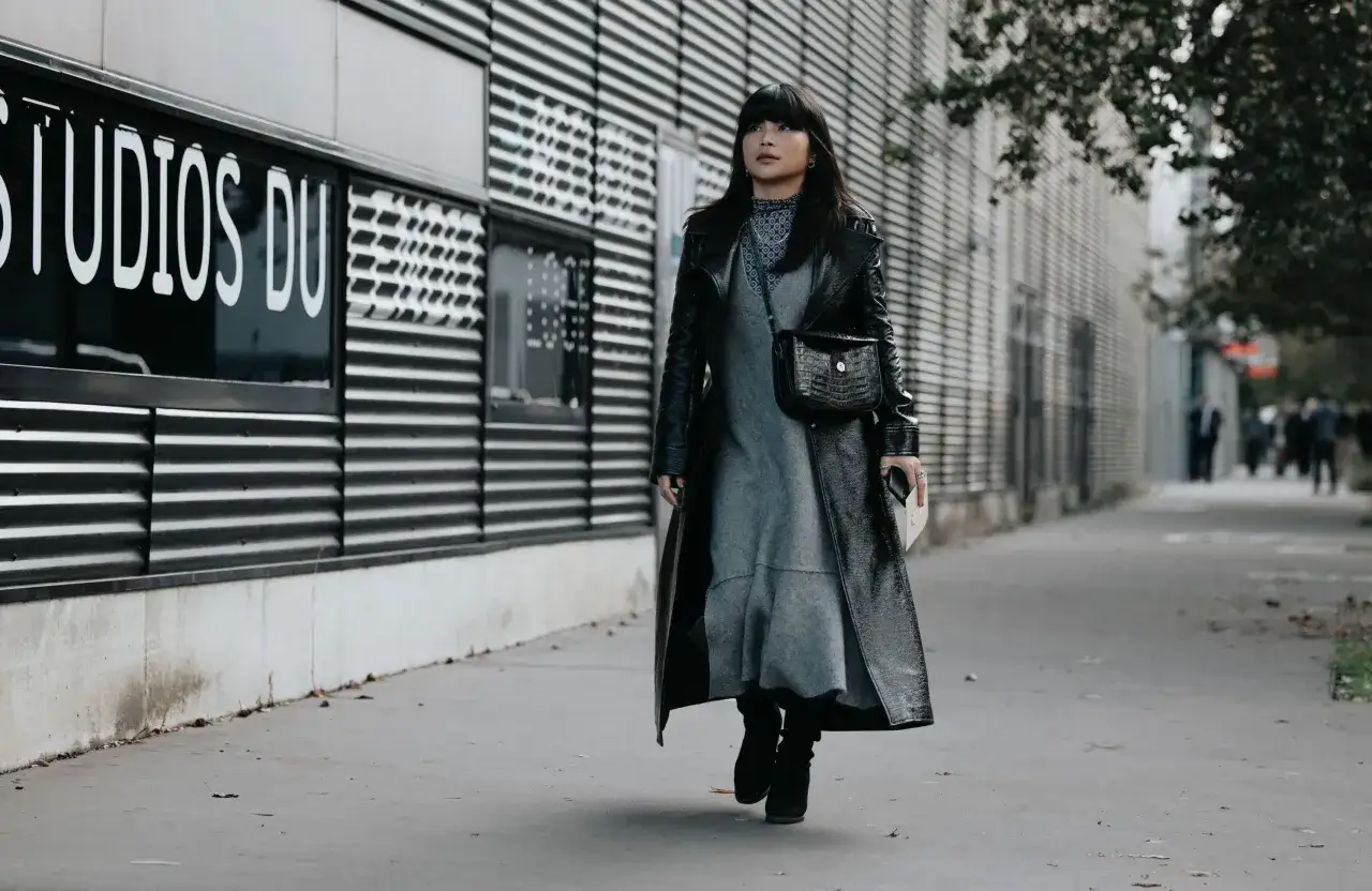 Une femme porte une robe noire et un long manteau en cuir verni, avec des bottines noires. Quelle chaussure avec une robe noire en hiver ? Des bottines, c'est parfait !