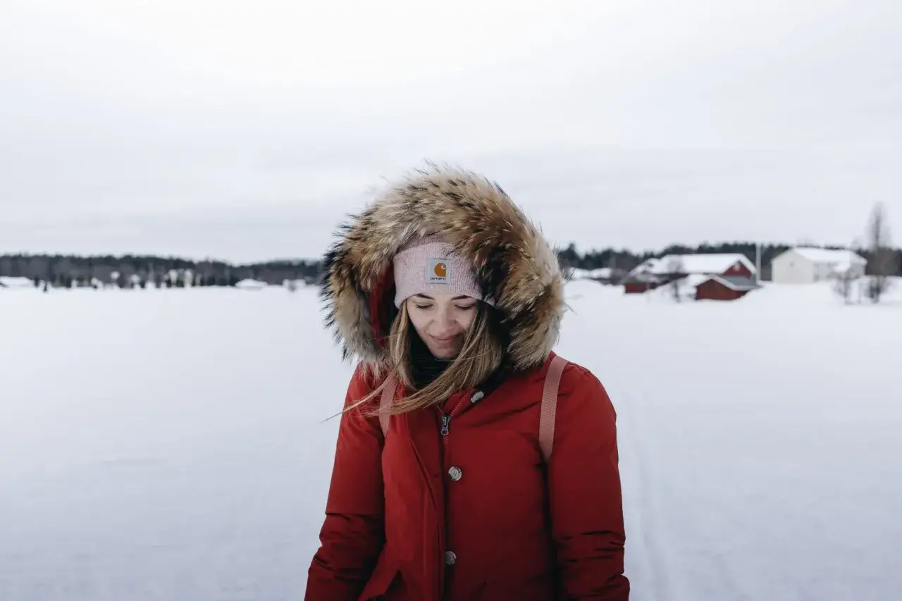 Jeune femme en parka rouge à capuche bordée de fourrure, bonnet rose, dans un paysage enneigé. Parfait pour savoir comment s'habiller pour ne pas avoir froid.