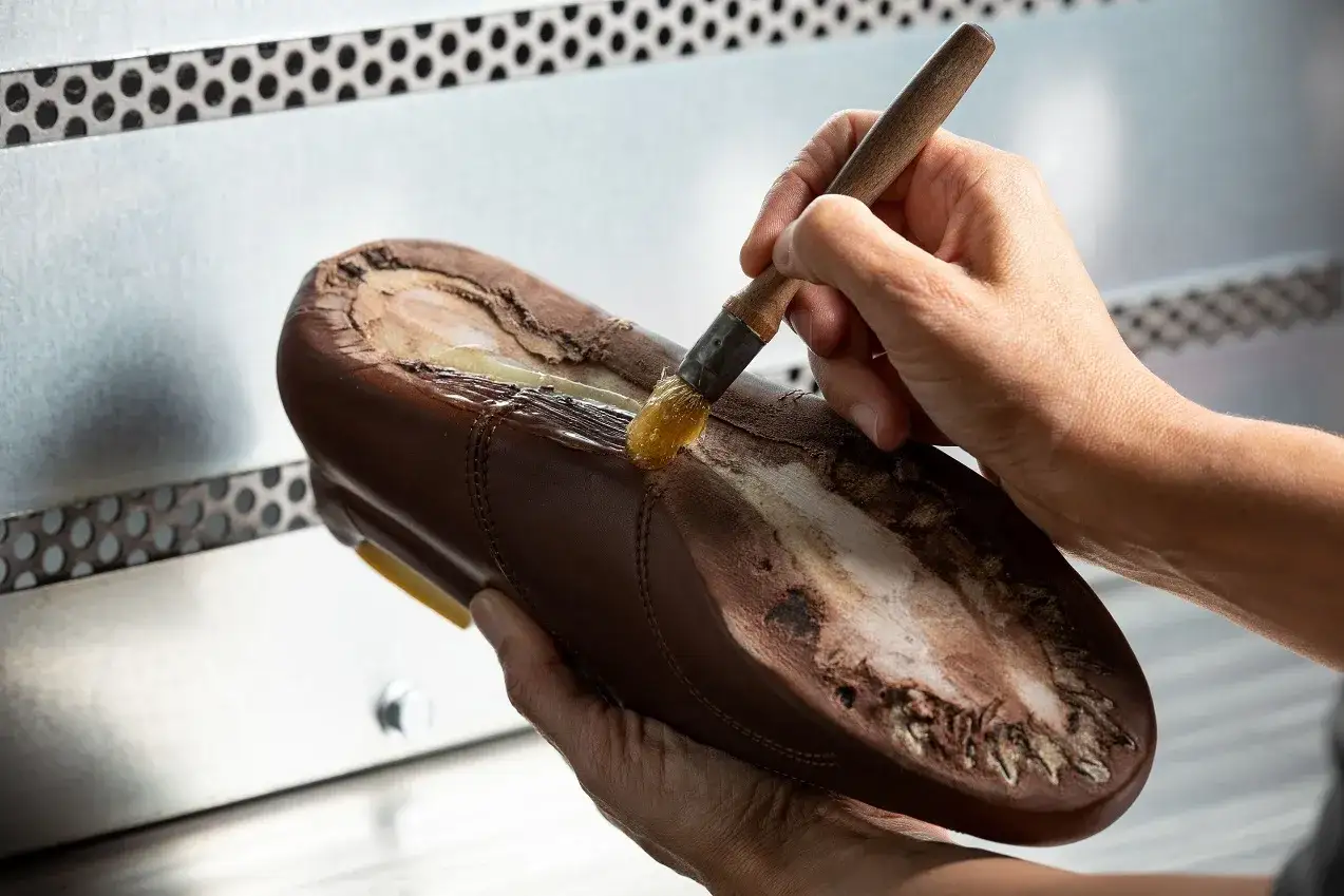 Main d'artisan appliquant de la colle sur une semelle de chaussure marron pour la réparer. L'artisan utilise un pinceau pour poser soi même patin chaussure.