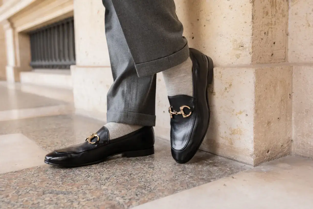 Un homme porte un costume gris avec des mocassins noirs ornés d'une boucle dorée. Parfait pour un look élégant.