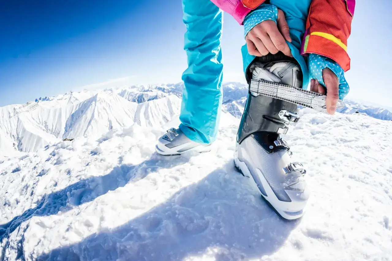 Préparation pour dévaler les pistes : une personne ajuste sa chaussure pour aller au ski, entourée de montagnes enneigées sous un ciel bleu éclatant.