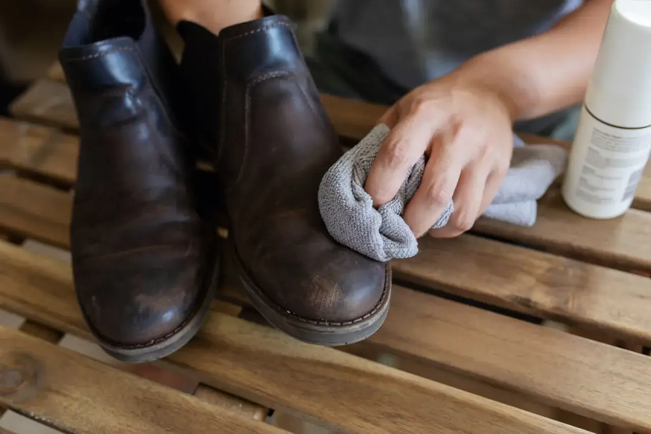 Entretien simili cuir : une main nettoie une botte marron avec un chiffon gris. Un produit d'entretien est à côté.