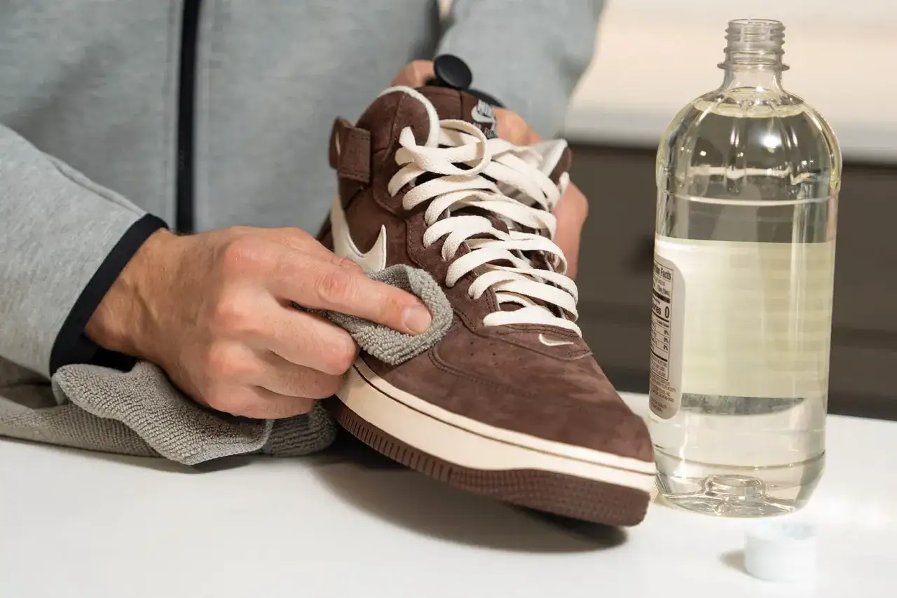 Une astuce de grand-mère pour nettoyer le daim : une main frotte délicatement une chaussure marron avec un chiffon doux, à côté d'une bouteille de liquide clair.