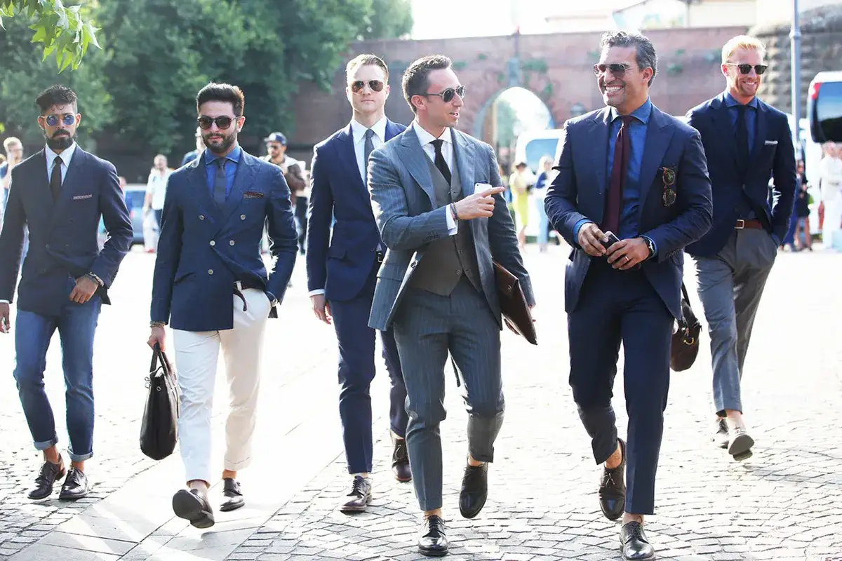 Un groupe d'hommes élégants marchent, portant chacun un costume sans chaussette. Leurs tenues varient, allant du bleu marine au gris rayé, avec des détails soignés.