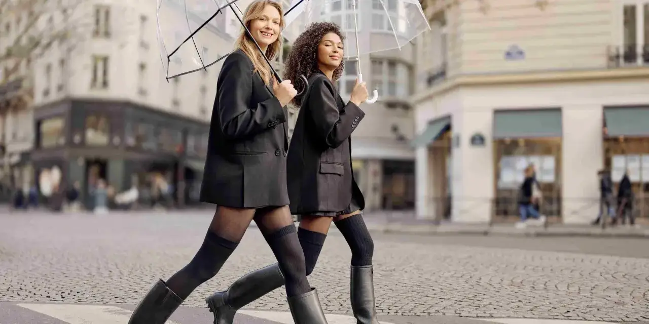 Deux femmes traversent une rue pavée sous des parapluies transparents, portant des blazers noirs et des tenues avec collant noir opaque et bottes.