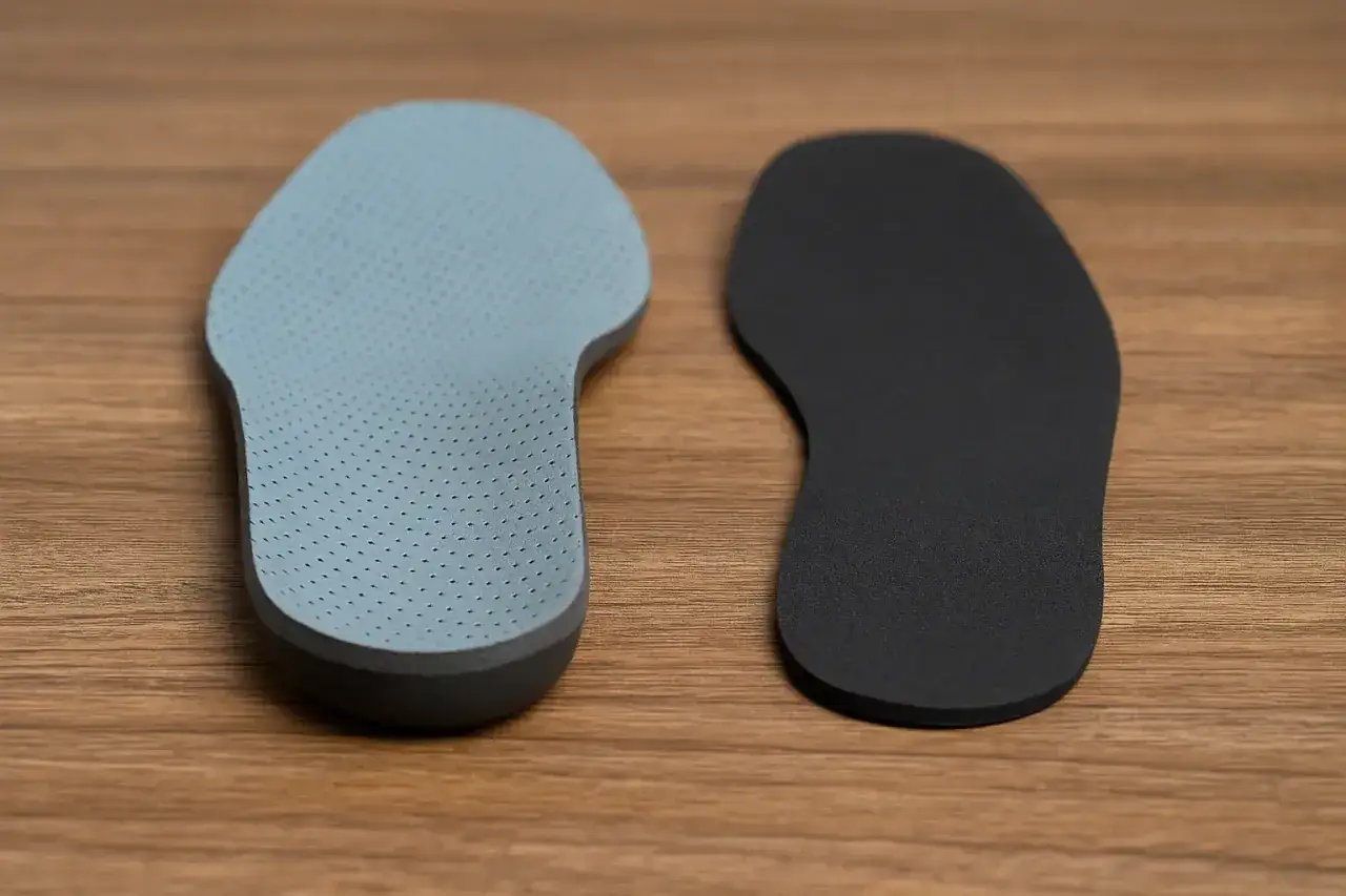 Deux semelles orthopédiques, une grise perforée et une noire lisse, sur fond de bois. Avis semelle élastomère.