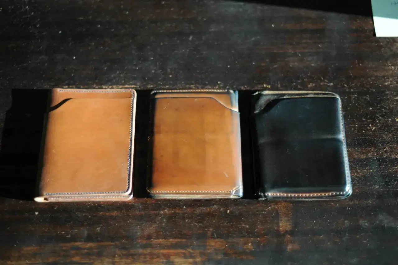 Trois portefeuilles en cuir Horween, un clair, un brun et un noir, posés sur une table en bois sombre.