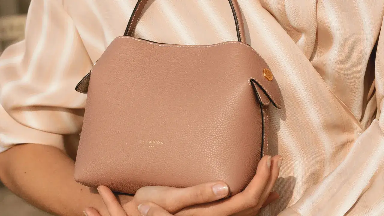 Sac rose poudré en cuir grainé, tenu par des mains délicates, sur une blouse à rayures beiges.