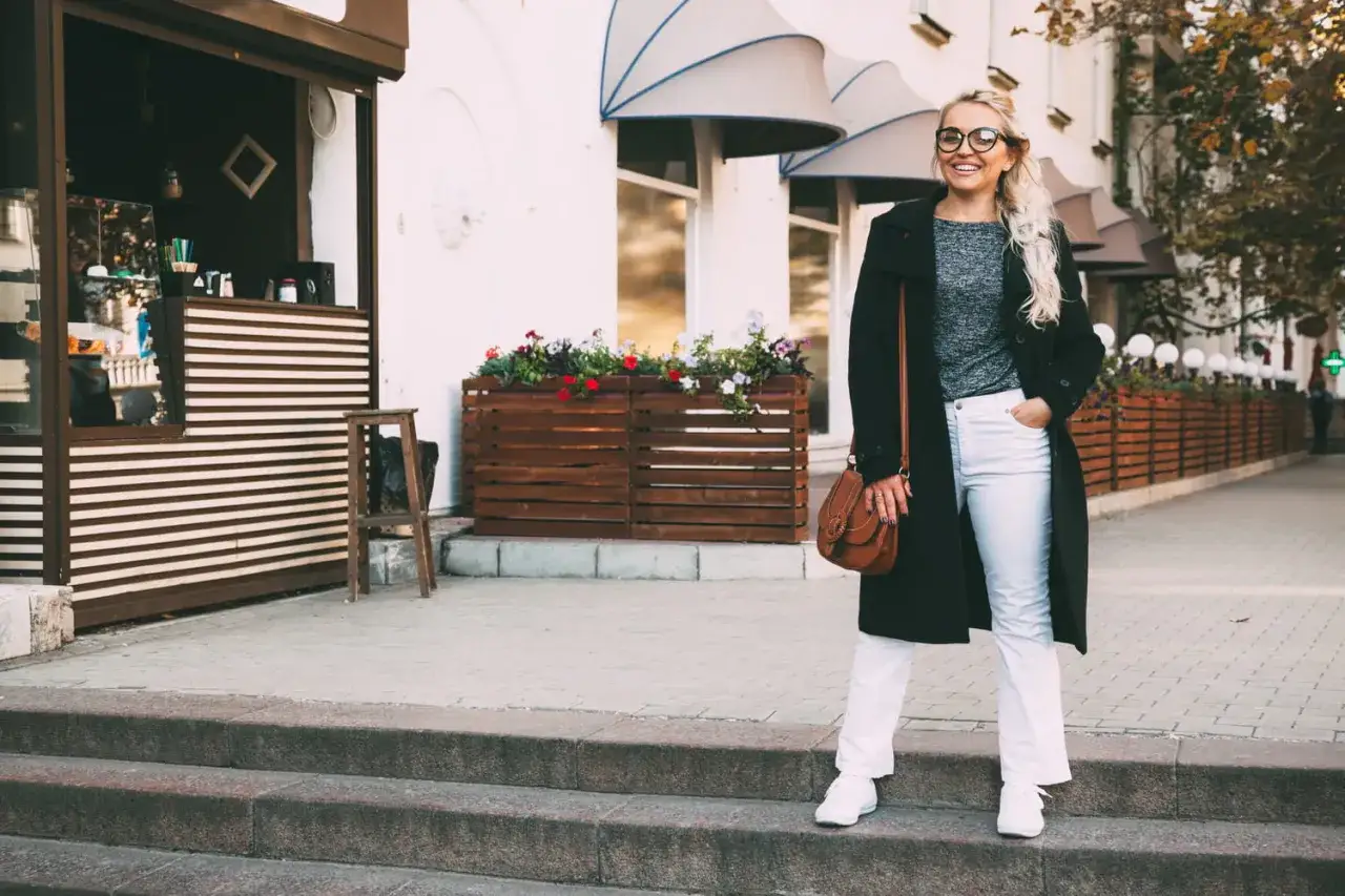 Femme souriante en jean bootcut, manteau noir, sac marron et baskets blanches, devant un kiosque.