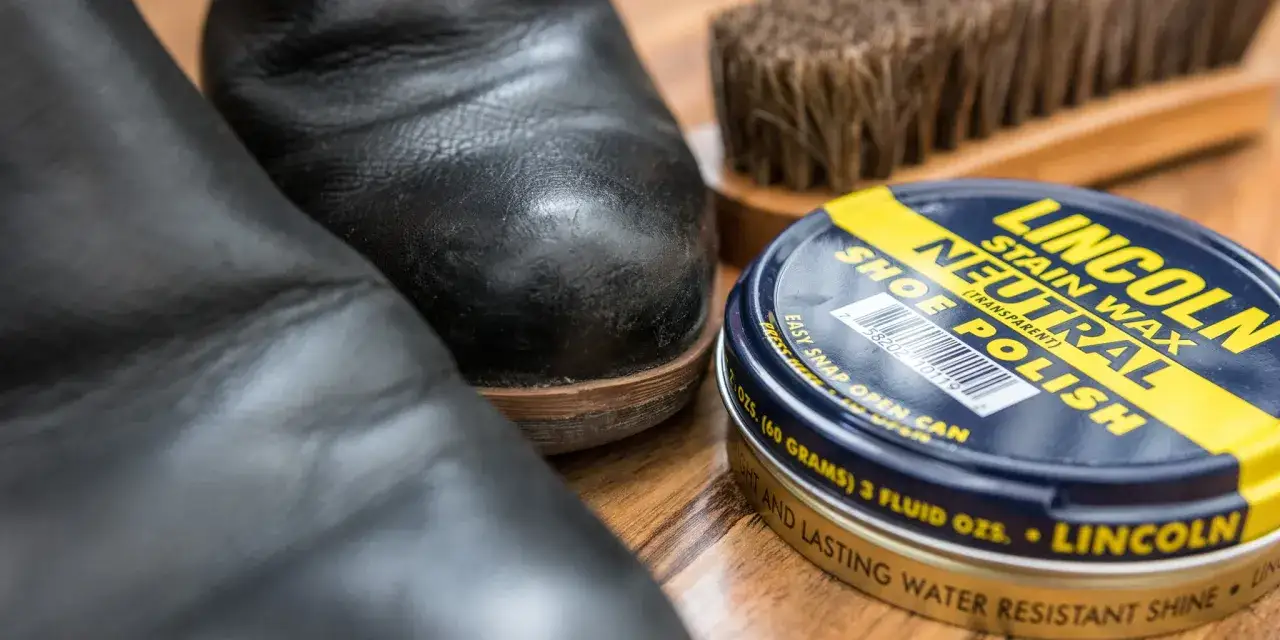Deux bottes noires, une boîte de cirage sec Lincoln et une brosse à chaussures sur fond de bois.
