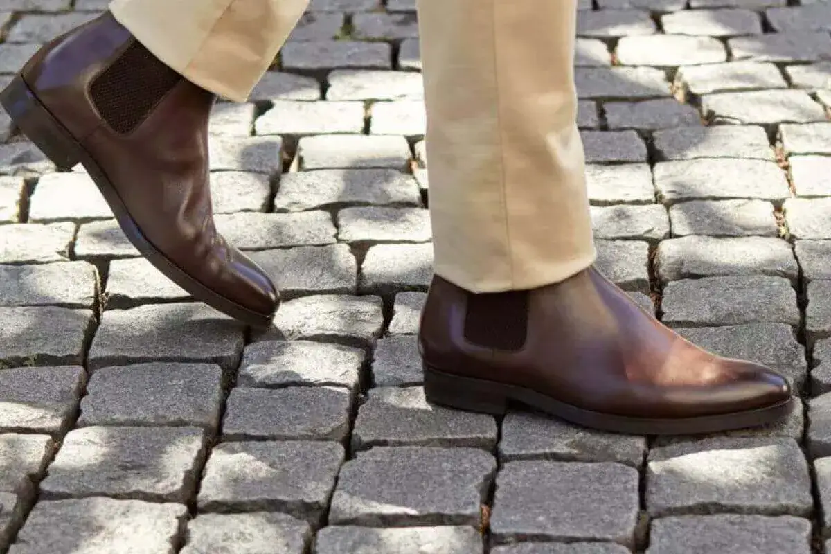 Un homme marche sur des pavés, ses bottines marron à semelle cuir ou gomme brillent sous le soleil.