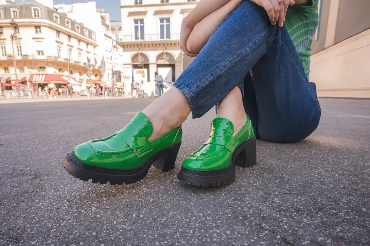 Chaussures vertes en cuir vernis, avec une plateforme épaisse, portées par une personne assise dans la rue, avec des bâtiments parisiens en arrière-plan.